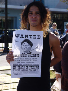 Youth holding sign that reads "WANTED CHRISTOPHER COLUMBUS - GRAND THEFT, GENOCIDE, RACISM, INITIATING THE DESTRUCTION OF A CULTURE, RAPE, TORTURE, AND MAIMING OF INDIGENOUS PEOPLE AND INSTIGATOR OF THE BIG LIE. 500 YEARS OF TOURISM. A NATIVE AMERICAN PERSPECTIVE. STEAL THIS POSTER - YOU'VE STOLEN EVERYTHING ELSE"