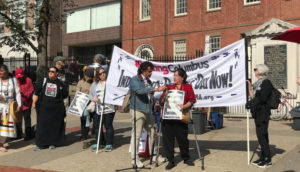 Speaker using mobility devices in front of banner that reads #SinkingColumbus Indigenous Peoples' Day Now!