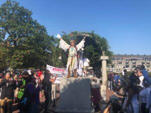 Chali'Naru Dones, Taíno, speaking with arms outstretched to a crowd from on top of the column that formerly held the Columbus Day Statue in Boston